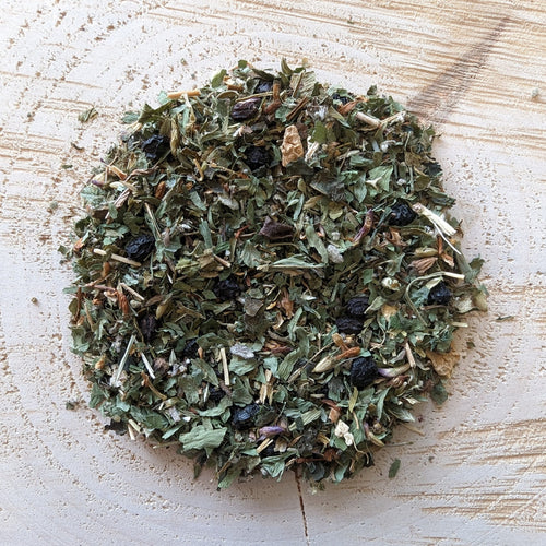 A mixture of dried herbs and leaves, including nettle, elderberries, red clover, St. John's Wort, sage, ginger root, and alfalfa, arranged on a wooden surface.
