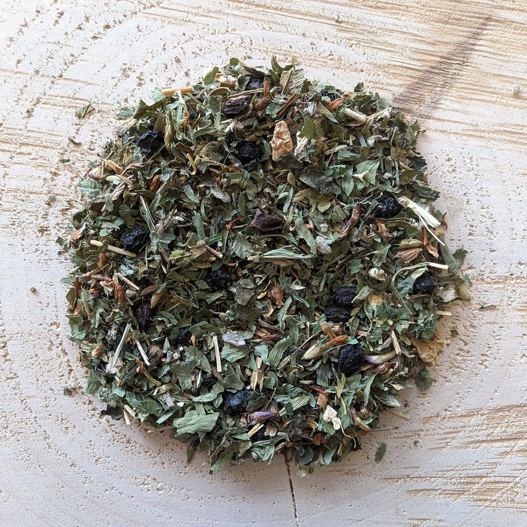 A mixture of dried herbs and leaves, including nettle, elderberries, red clover, St. John's Wort, sage, ginger root, and alfalfa, arranged on a wooden surface.